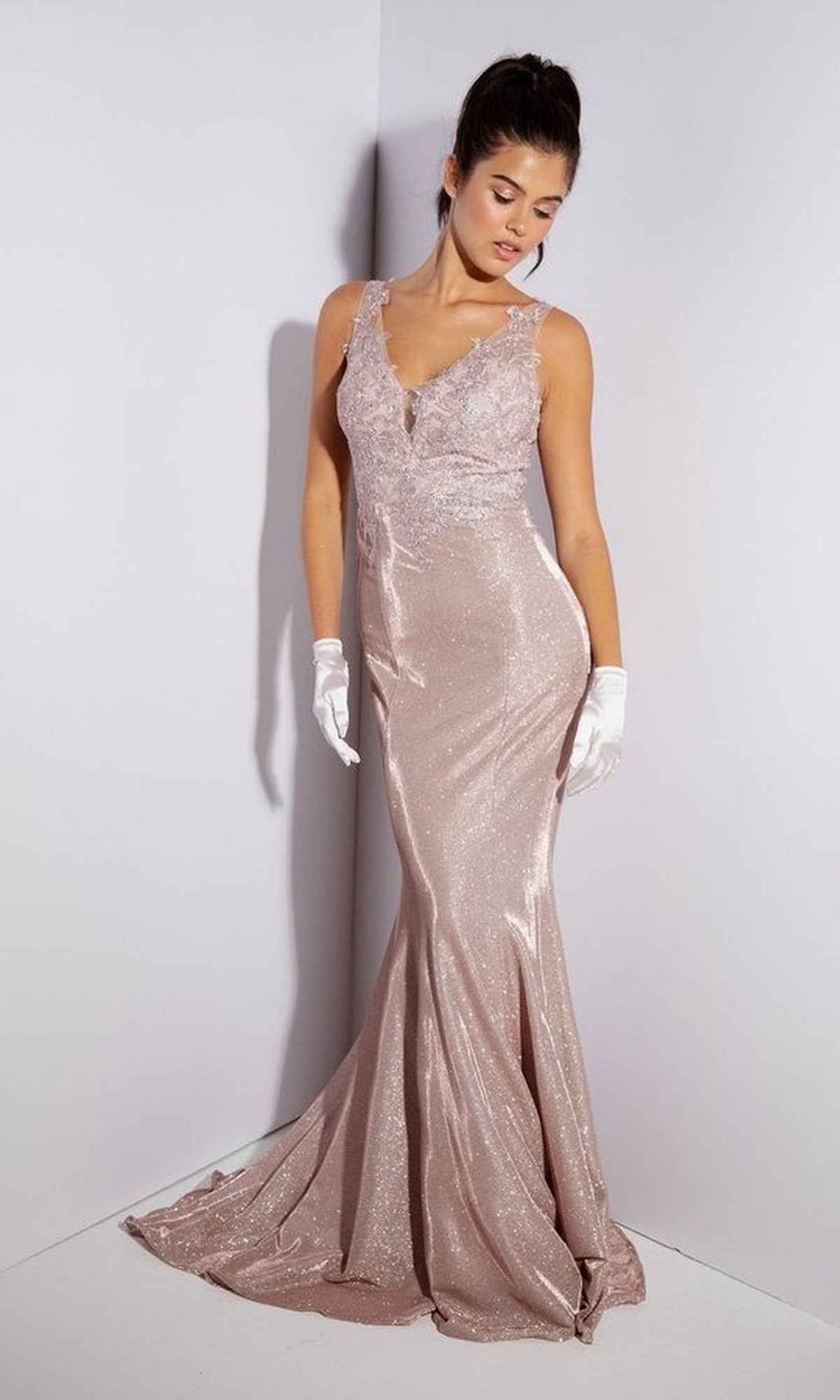 A woman wears the Eureka 9910 long prom dress featuring a shimmering light pink, floor-length skirt and lace-embroidered bodice with white gloves, standing against a plain white corner background.